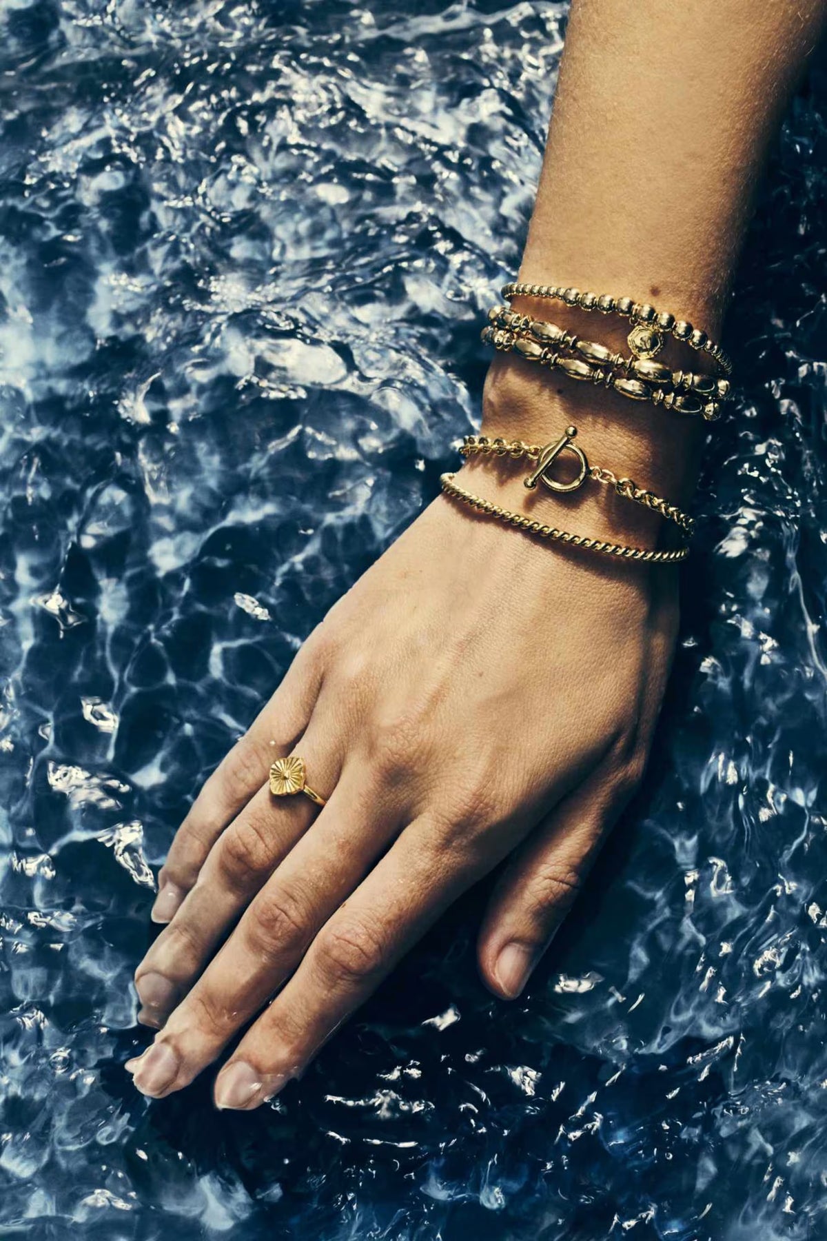 A close-up of a person's hand resting in deep blue water, adorned with multiple gold bracelets on the wrist and a gold ring on the ring finger.