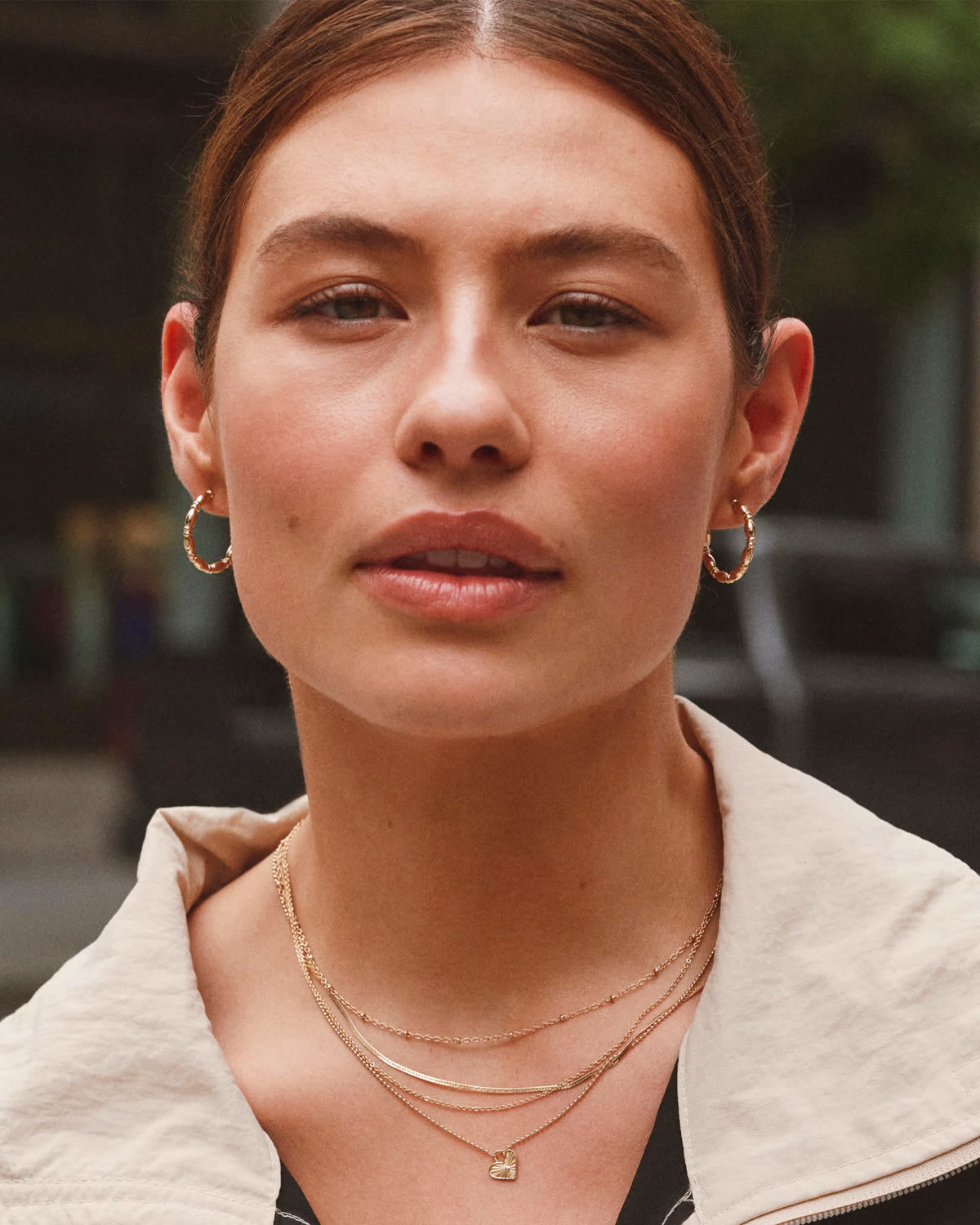 A close-up portrait of a woman wearing layered gold necklaces, hoop earrings, and a small gold box pendant, with a blurred outdoor background.