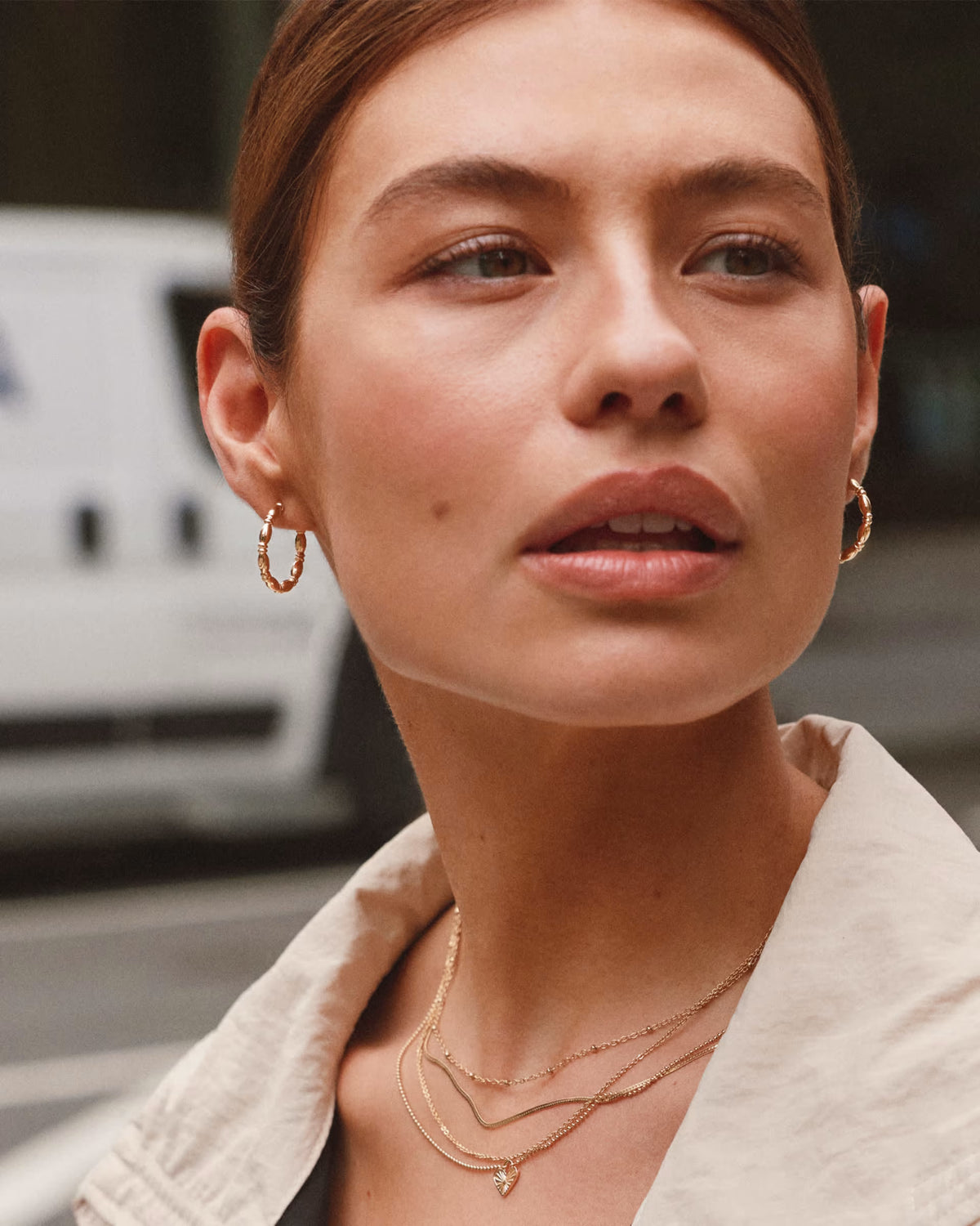 A close-up lifestyle shot of a woman wearing gold jewelry, including a small gold hoop earring and layered gold necklaces, with a blurred outdoor background.
