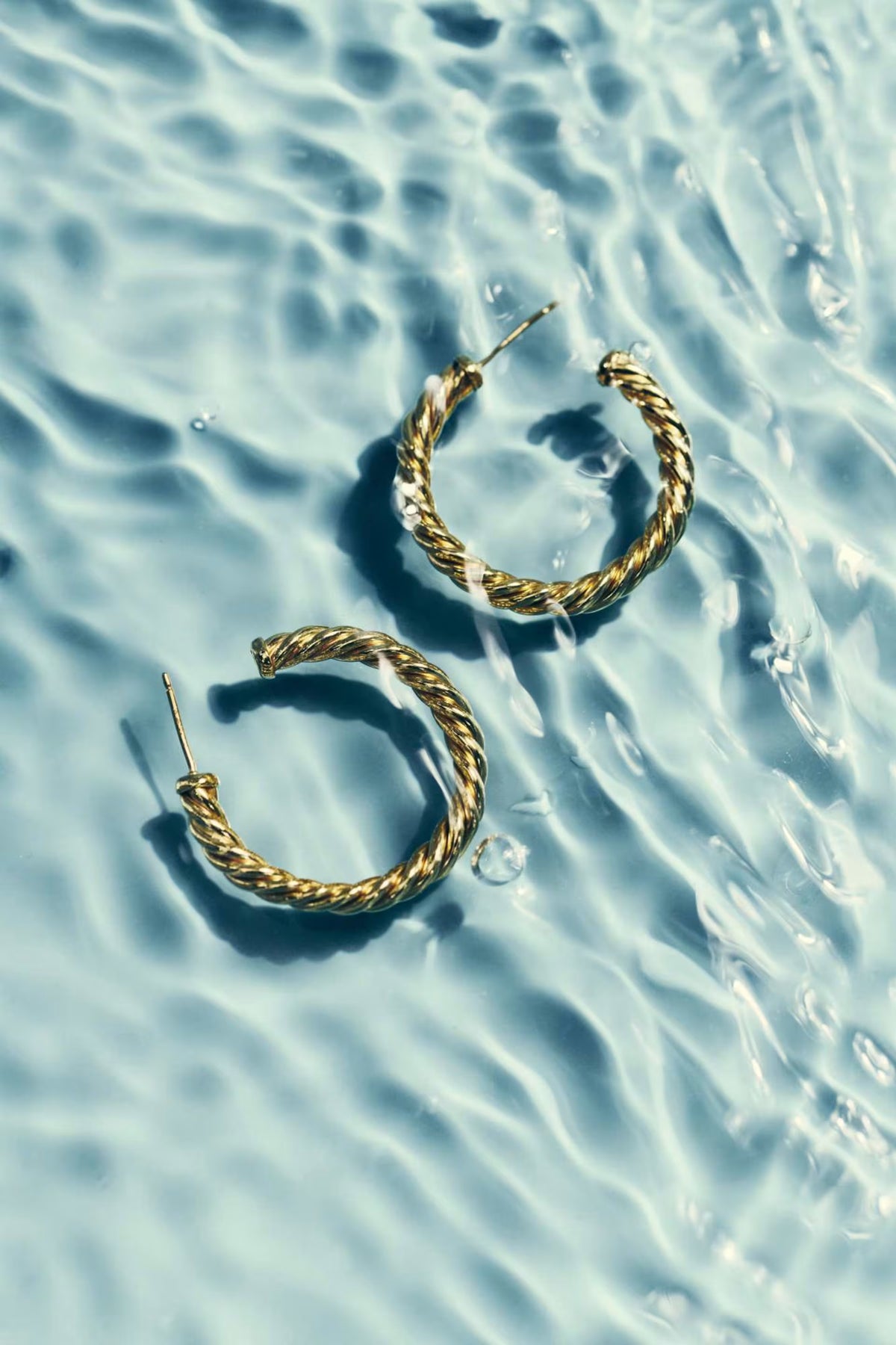 A pair of gold twisted hoop earrings resting on rippling light blue water.