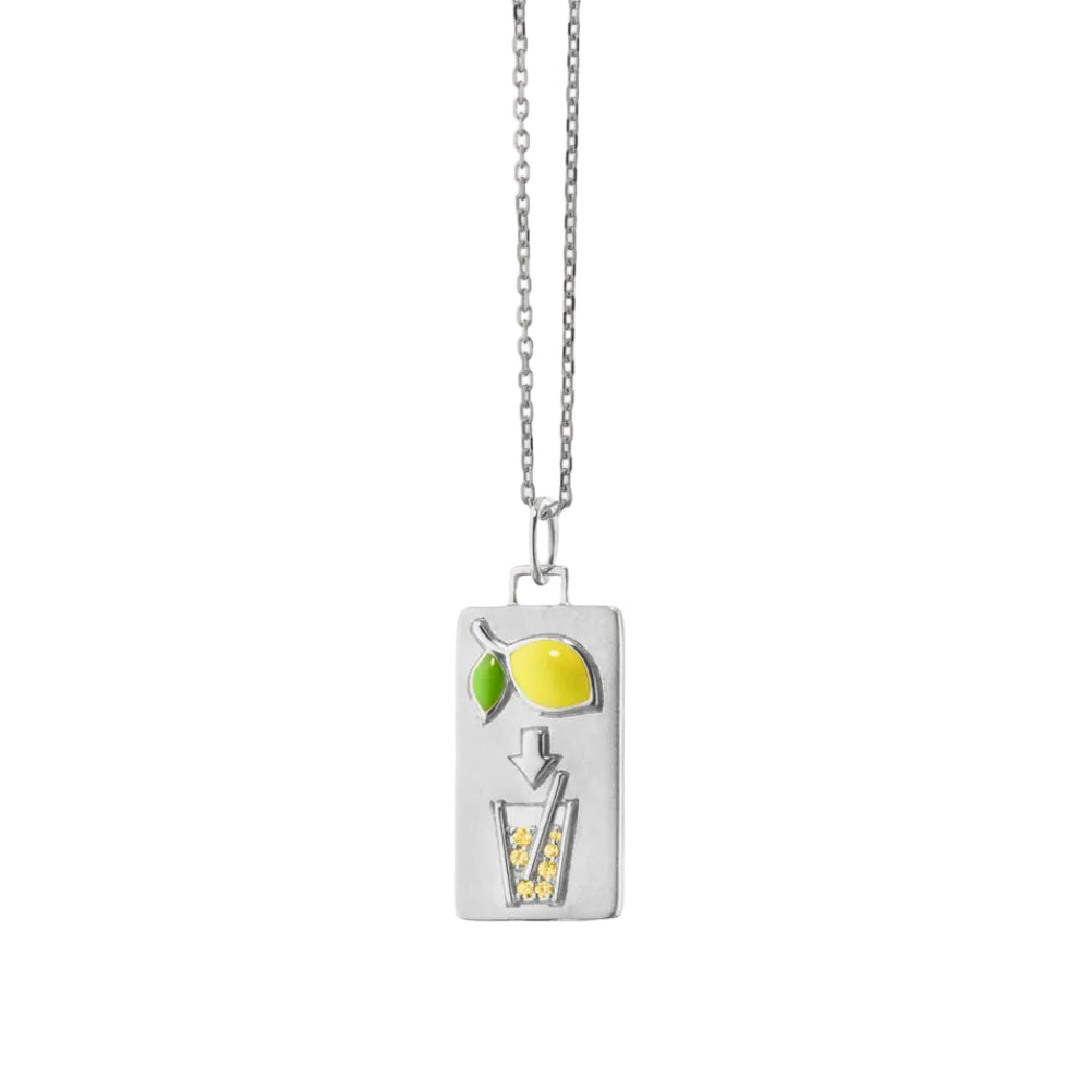 A silver rectangular pendant necklace featuring a lemon-themed illustration with a yellow lemon, green leaf, downward arrow, and a glass of lemonade filled with small yellow dots, shown on a light cream background.
