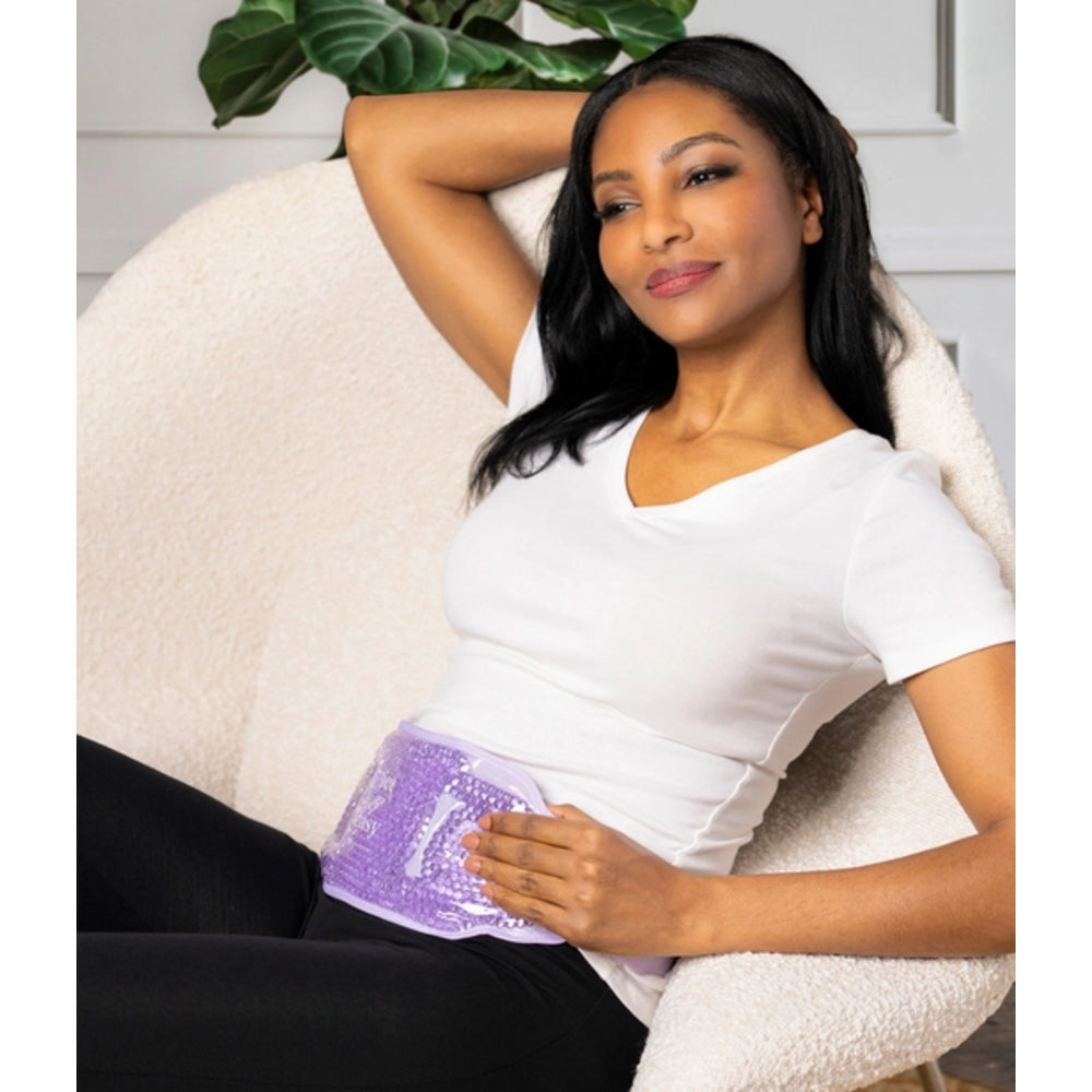 A woman reclining in a cream-colored armchair, wearing a white t-shirt and black leggings while demonstrating a purple and white textured abdominal ice pack on her lower stomach.