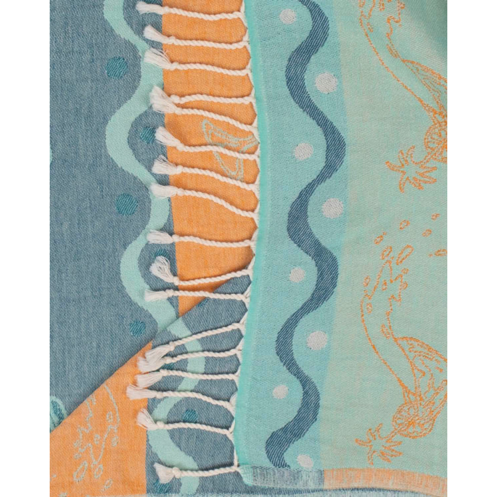 A close up of a blue beach towel with beach-themed design featuring martini glasses and wavy lines and white tessal edges on a blue and orange background