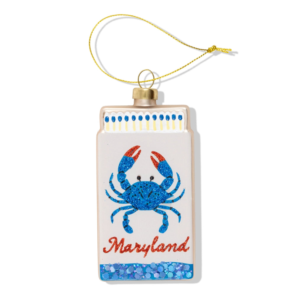 A rectangular Christmas ornament designed to look like a matchbox, featuring a glittery blue crab with red claws and the word 'Maryland' in red script. The ornament has a gold hanging string and is set against a plain white background.