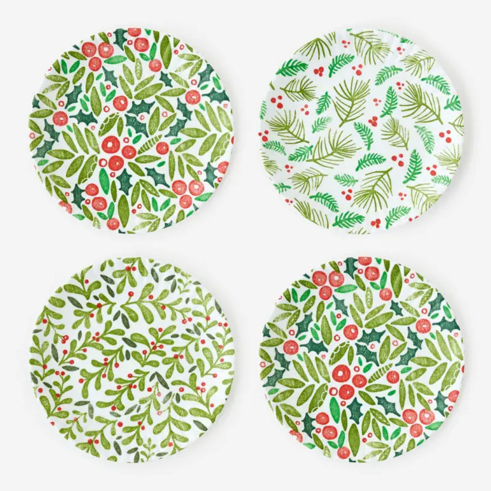 Four round white plates arranged in a 2x2 grid, each decorated with a different holiday botanical pattern featuring green leaves, red berries, and holly in watercolor style.