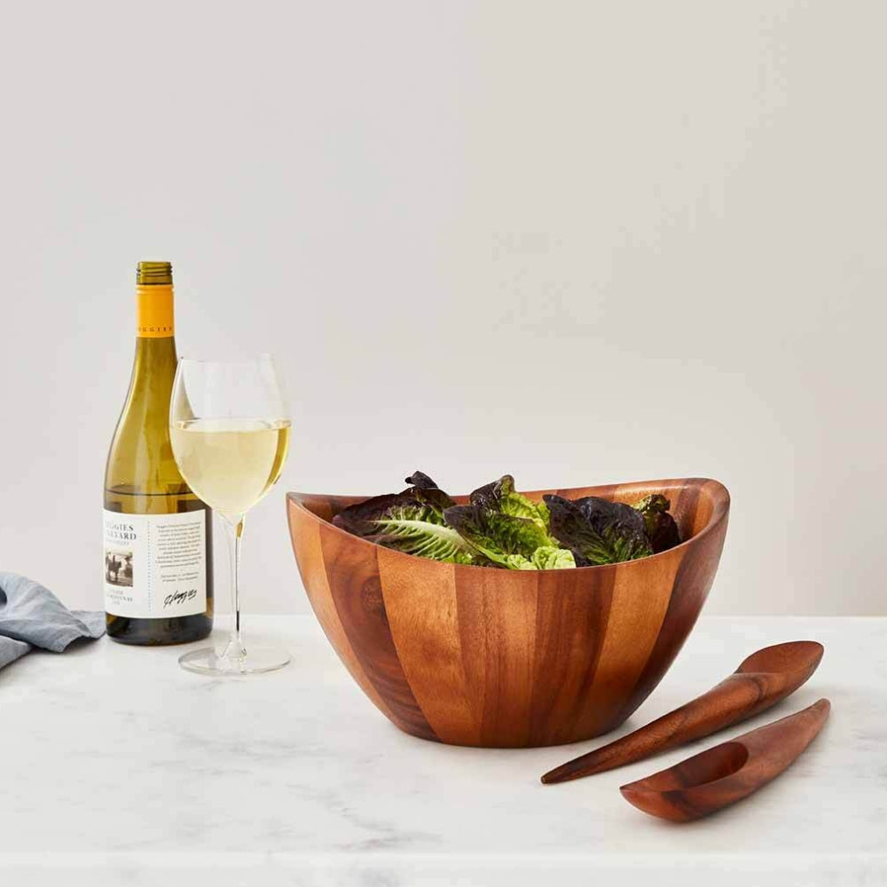 Wooden bowl and two wooden spoons with salad in the bowl next to a wine bottle and glass on a white table with a grey backround 