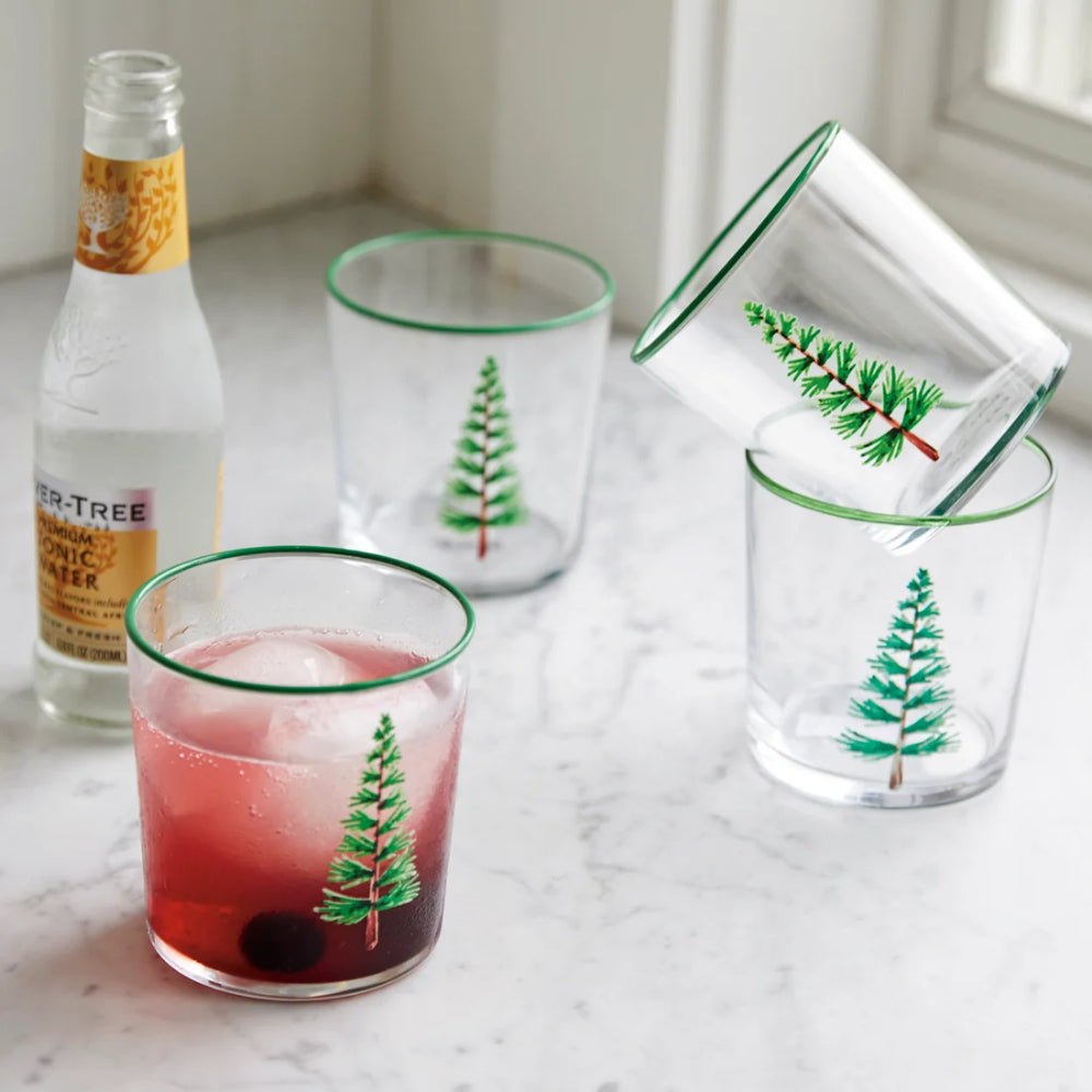 Trees double old fashioned glasses with a evergreen tree design an green rims on it on a with a glass bottle on a marbled white backround. 
