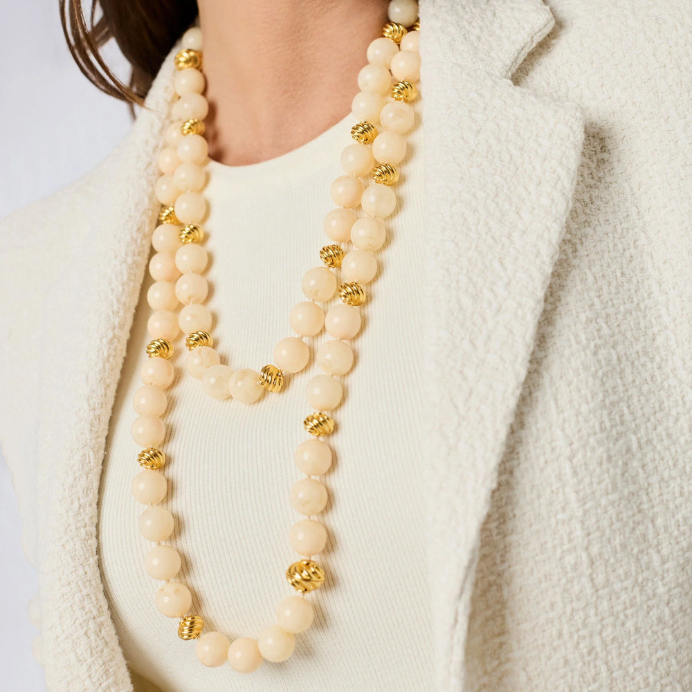 A close-up of a model wearing a double-strand cream-colored beaded necklace with textured gold spacer beads, styled with a cream knit top and a textured cream blazer.