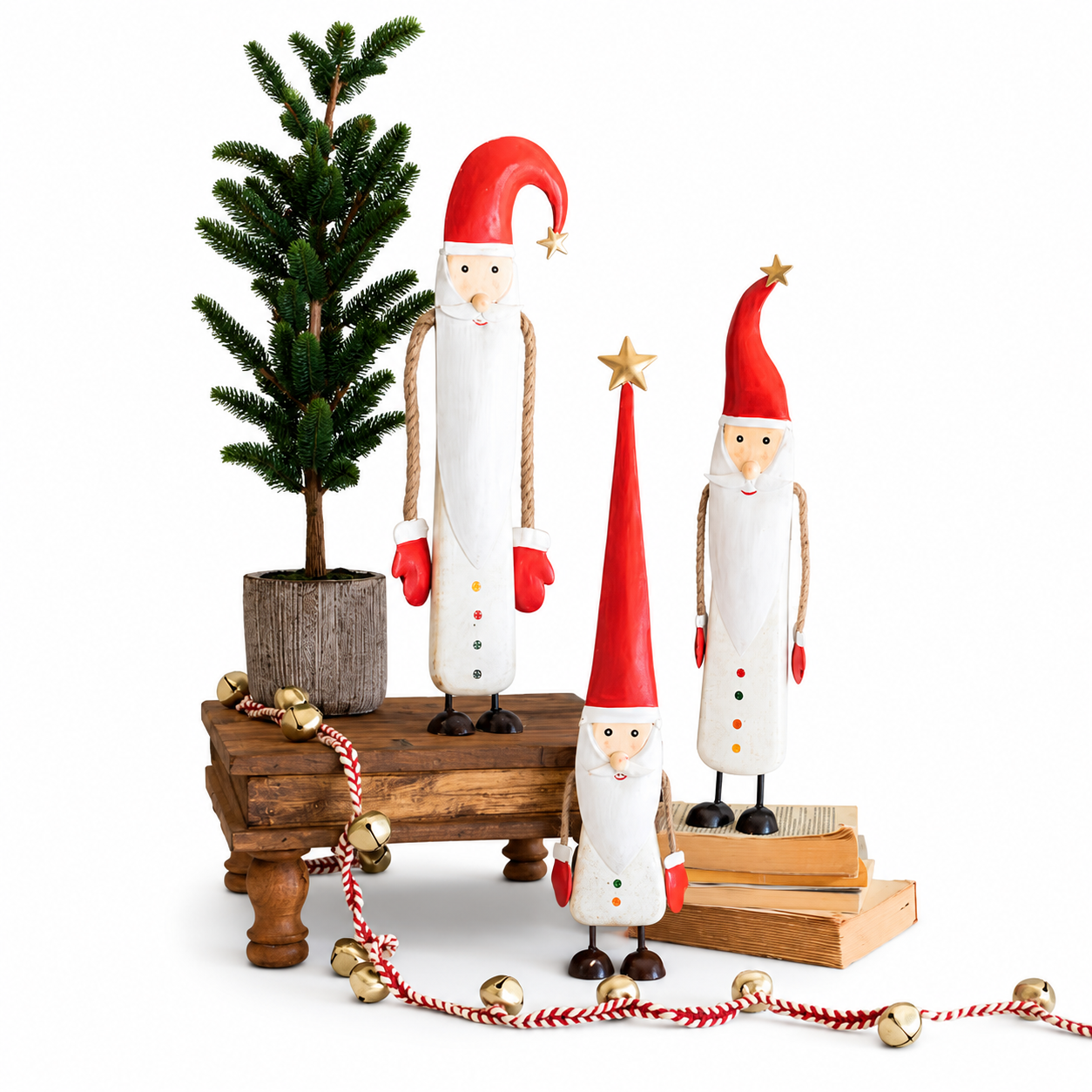 Three hand-painted wooden Christmas gnome figurines in white and red, styled on a rustic wooden bench with a small potted pine tree and red-and-white garland with gold bells against a white background.