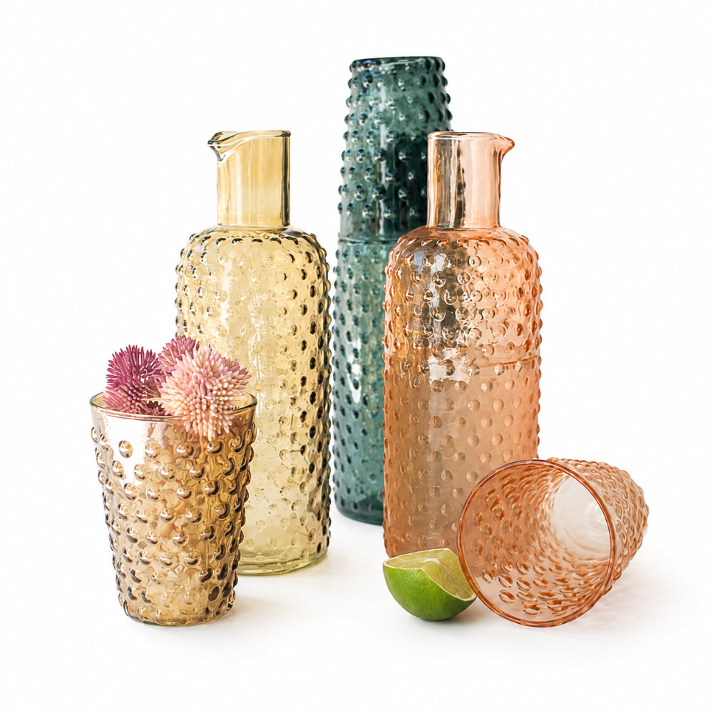 Three textured glass cocktail shakers in amber, teal, and copper tones, shown with matching textured tumblers and garnishes on a white background.