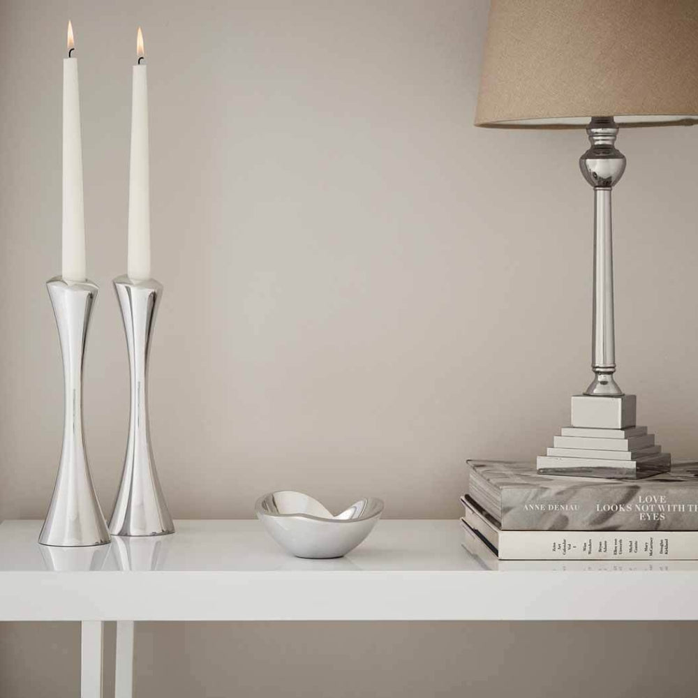 Decorative setup with silver candle holders, a bowl, books, and a lamp on a white surface.