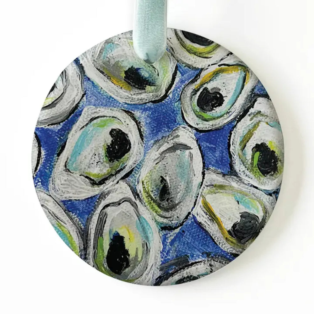 A round hanging ornament featuring an abstract painted pattern of oyster shells in blue, gray, white, green, and black, with a light blue ribbon loop at the top.
