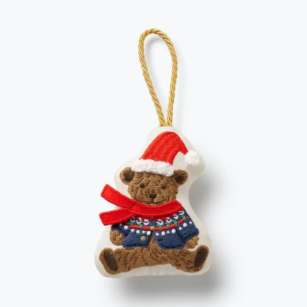 A plush teddy bear Christmas ornament with a brown body, wearing a red Santa hat with white trim and a red scarf with a blue and red patterned sweater, hanging from a gold-colored twisted cord on a white background.