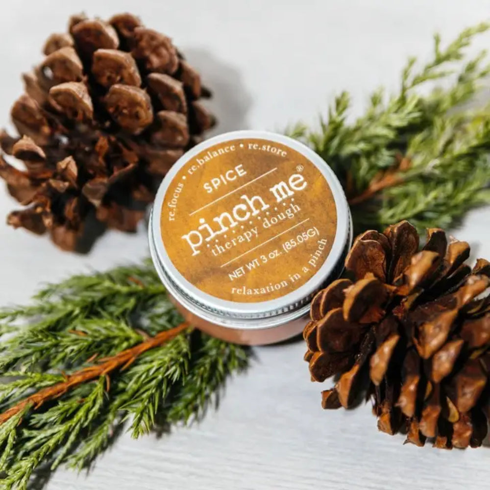 A Jar of Pinch Me Therapy Dough in flavor Spice nestled among pine cones and pine branches on a textured white background.