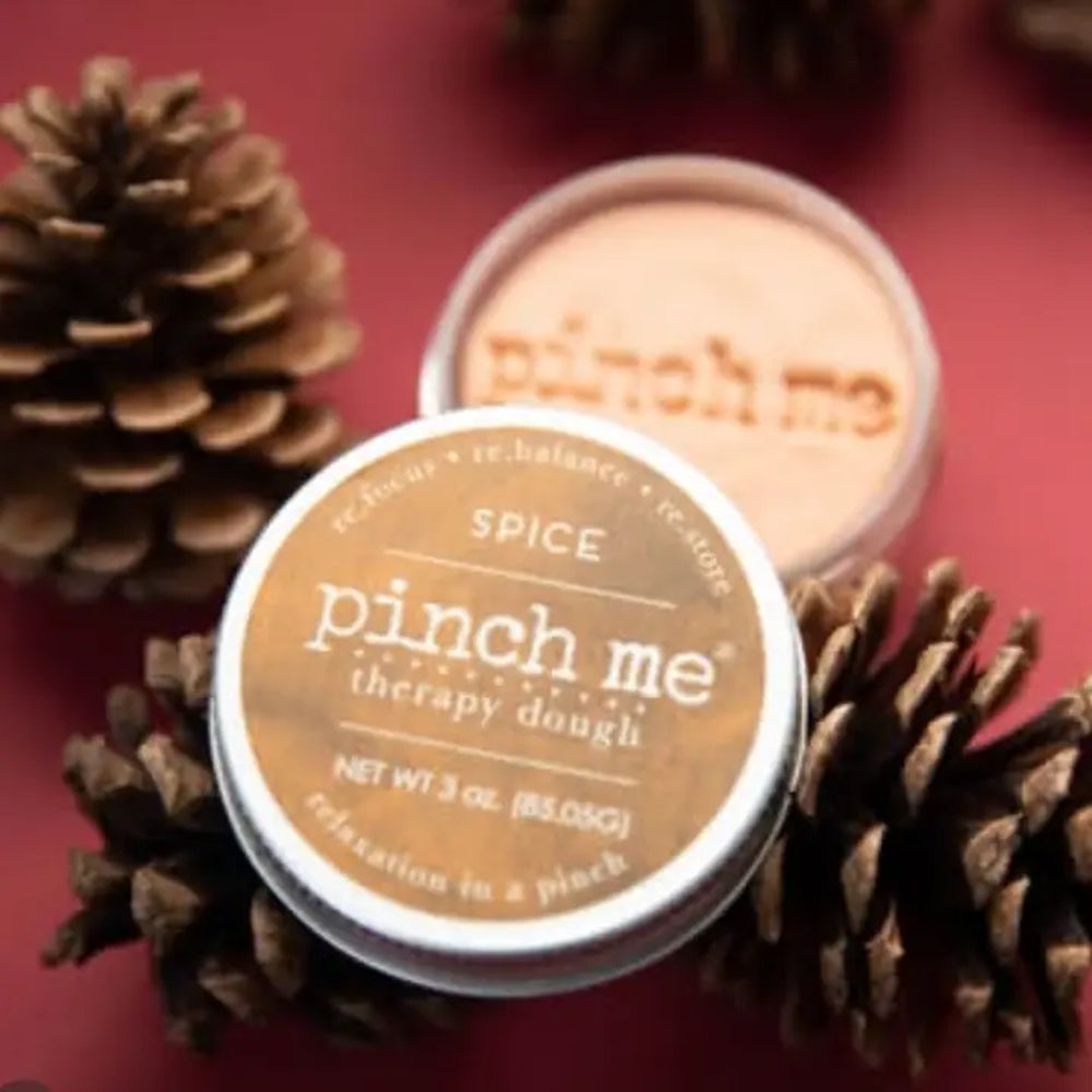 A jar of Pinch Me Therapy Dough in flavor Spice with the lid ajar to show the dough with the words "Pinch Me" pressed into it. Nestled among pine cones on a berry colored background.