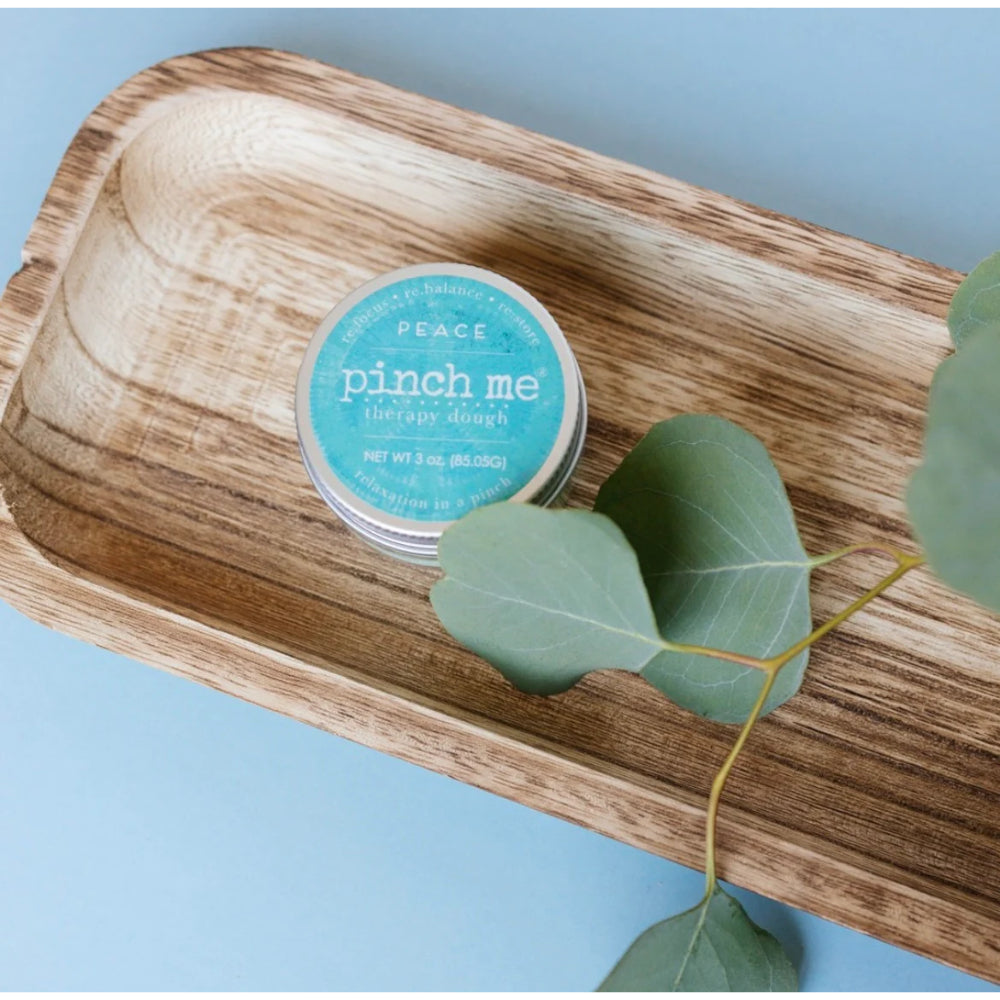 Jar of 'pinch me' therapy salve on a wooden tray with eucalyptus leaves against a light blue background.