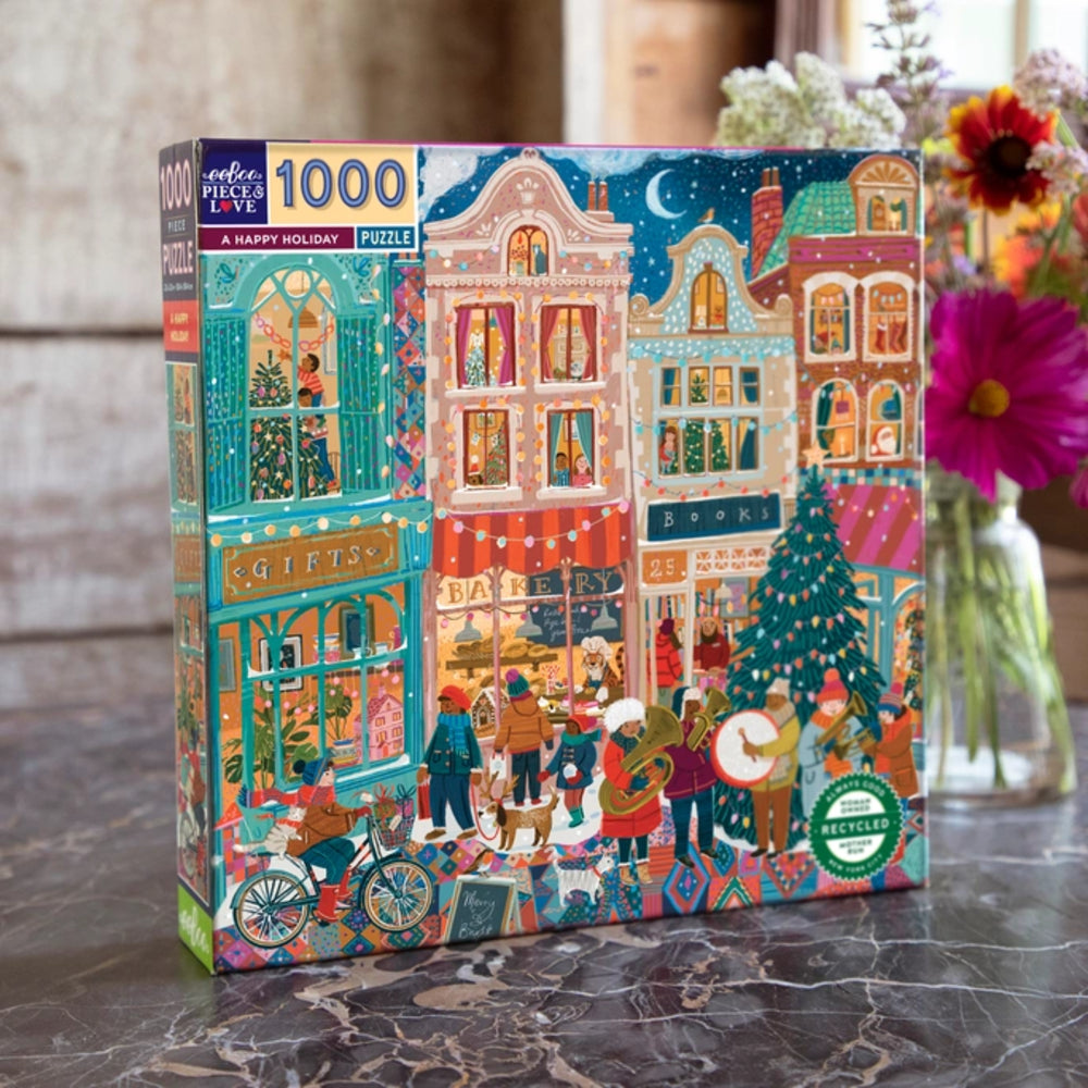 A 1000-piece jigsaw puzzle box titled 'A Happy Holiday' featuring a colorful, whimsical illustration of a Christmas town street with buildings, people, trees, and festive details. The box is displayed on a marble surface with flowers in the background.