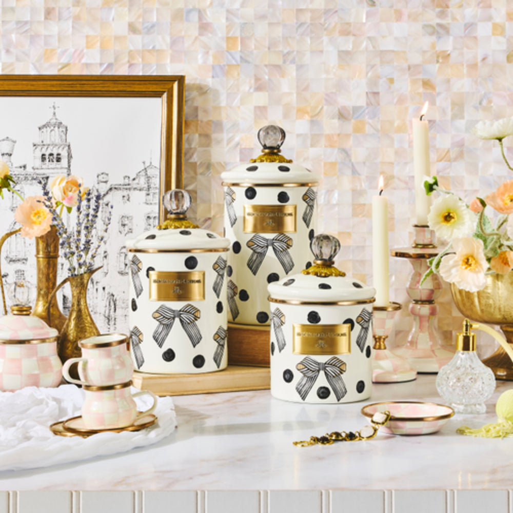 Three decorative enamel canisters with lids, featuring a white base, black polka dots, black bow patterns, and gold labels, styled on a marble countertop with matching pink and gold tableware in a kitchen setting.