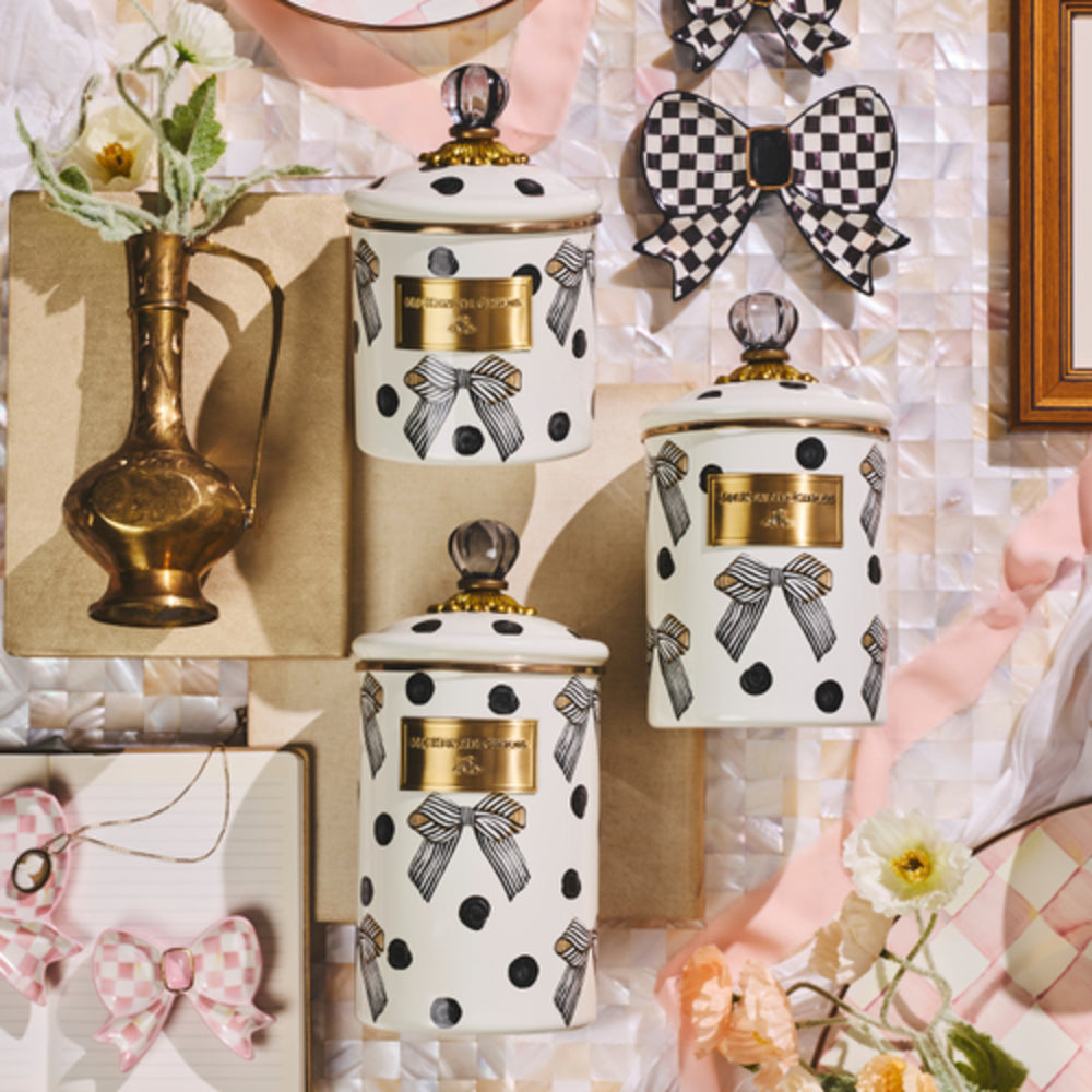 Three white enamel canisters with black polka dots, gray ribbon motifs, and gold labels, styled with pink ribbons, flowers, checkered bows, and a brass vase on a glossy shell backdrop.