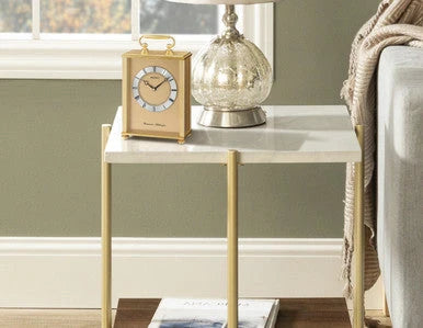 clock on a side table with lamp and window in a room with a couch and blanket