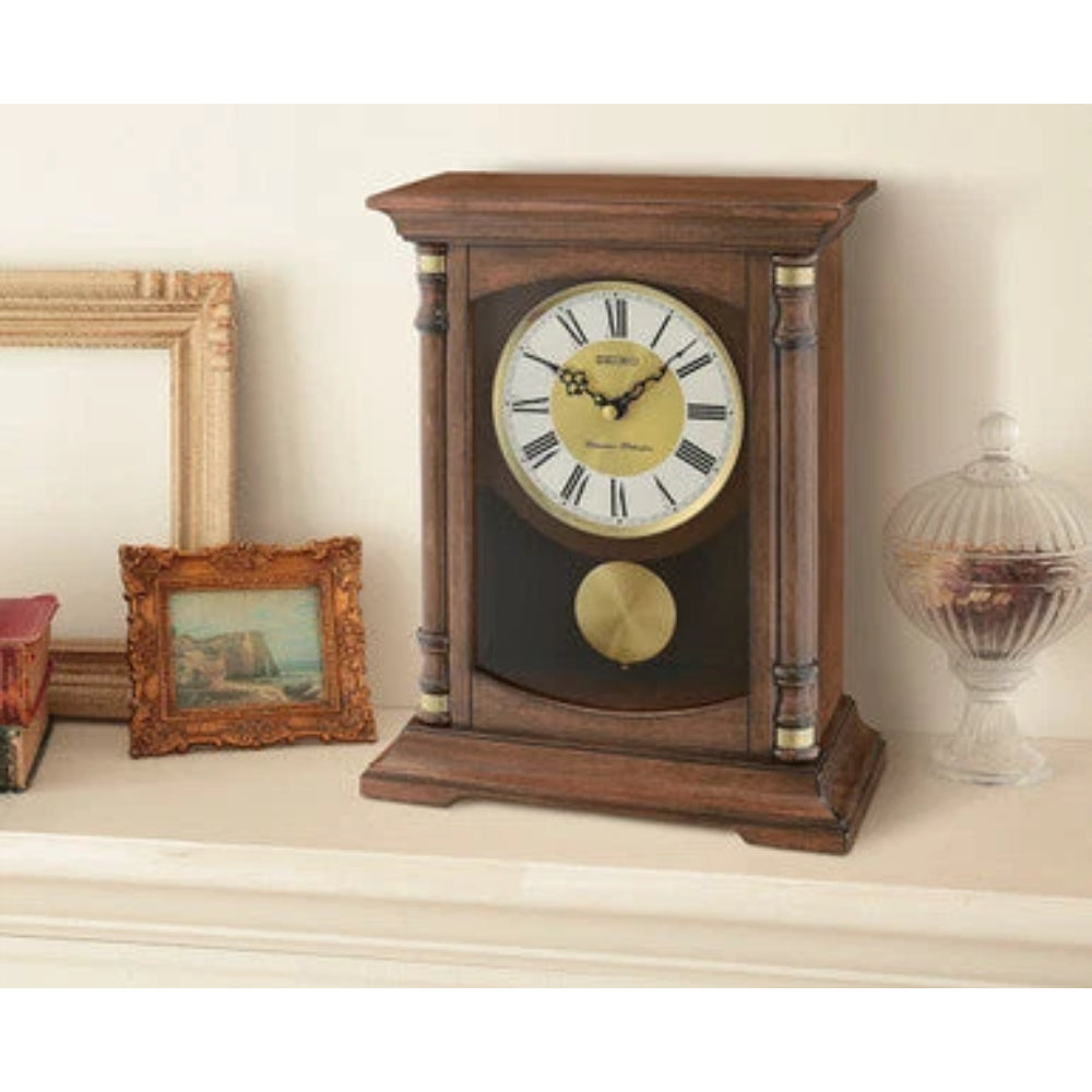 A traditional wooden mantel clock with a dark brown finish, a cream-colored dial with black Roman numerals, and a gold-toned pendulum, displayed on a shelf next to decorative frames and a glass jar.