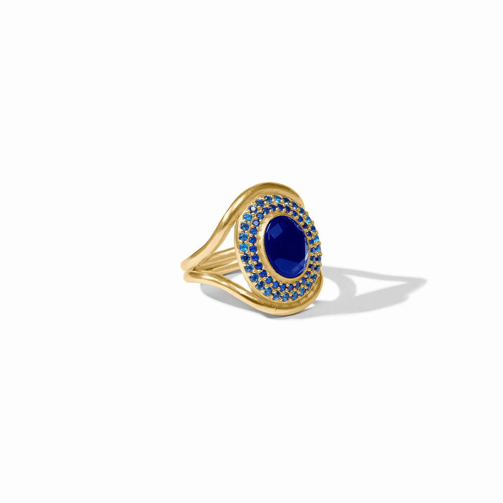 A gold cocktail ring with blue small stones surrounding a larger center blue stone on a white background