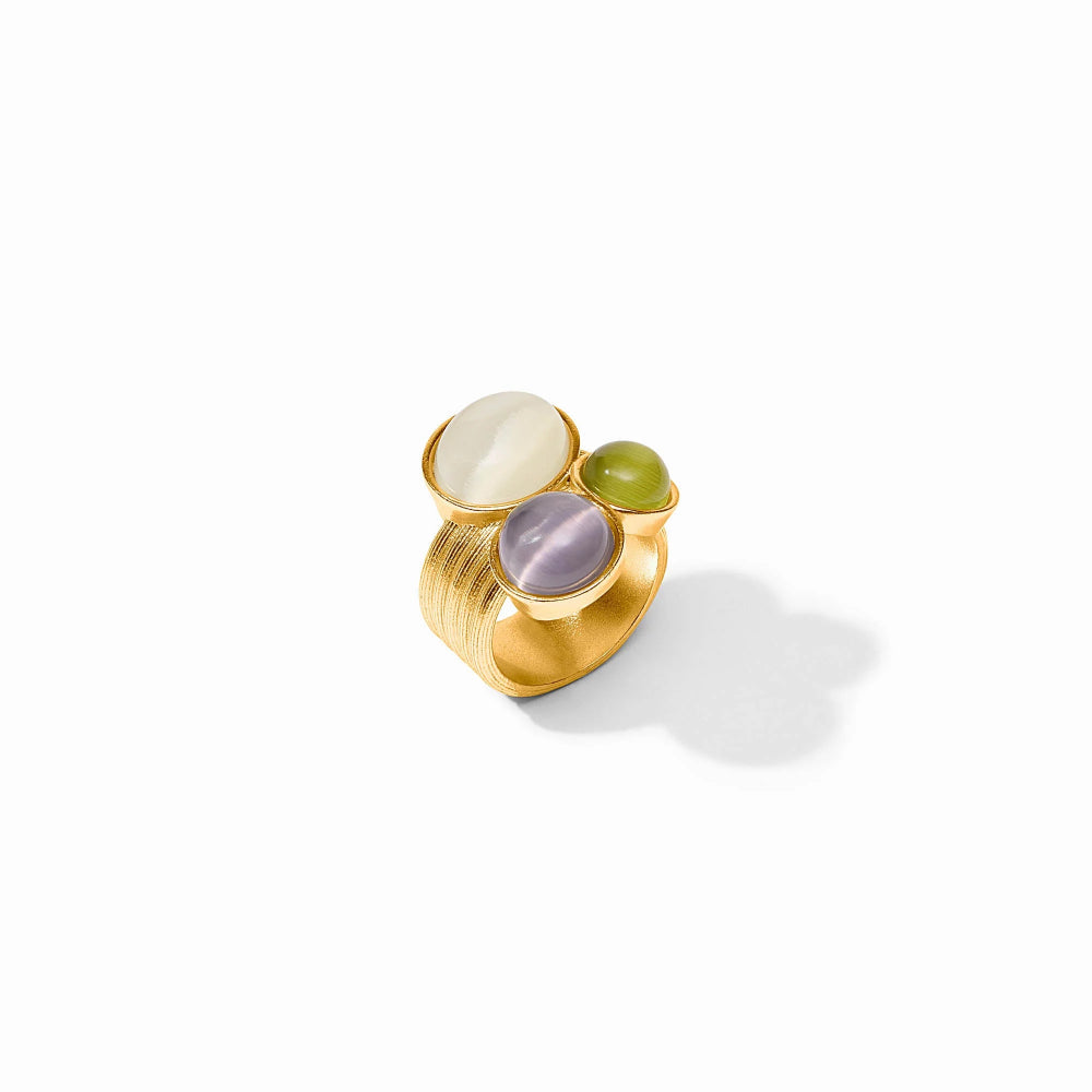 A gold ring featuring three oval gemstones in different colors: a cream cabochon, a light purple amethyst, and a green jasper, set against a clean white background with a soft shadow.