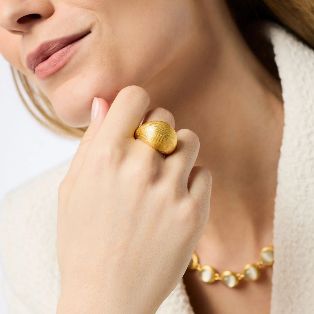 A close-up lifestyle shot of a model wearing a chunky, textured gold ring on her finger, with a matching gold necklace partially visible.