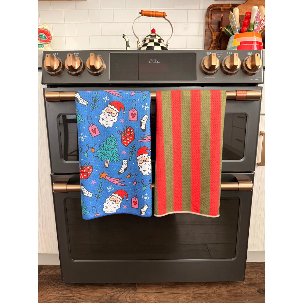 Two Christmas-themed kitchen towels hanging from the handlebars of a black gas range stove. The left towel is blue with a Santa Claus and holiday tree pattern, and the right towel has vertical red and green stripes.