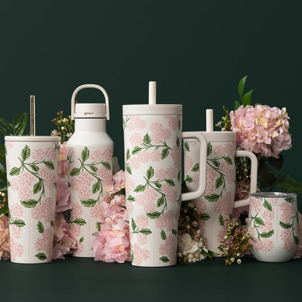 Pink cream colored stemless tumbler with a pink hydrangea floral design on a white background with other simular patterned tumblers and with flowers in the background on a dark green backround.