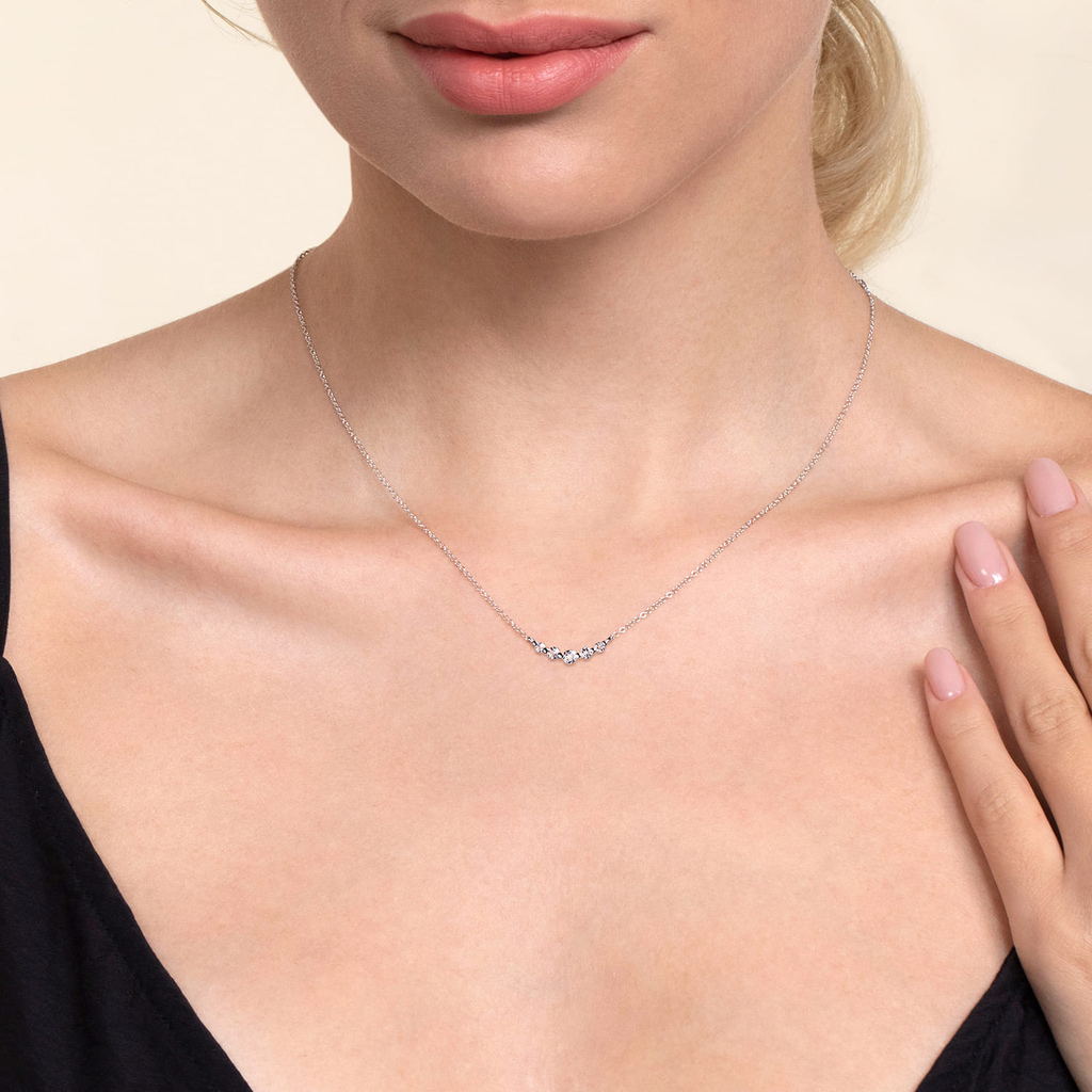 A close-up of a model wearing a delicate silver necklace with a small wavy pavé pendant, styled with a black top against a warm beige background.