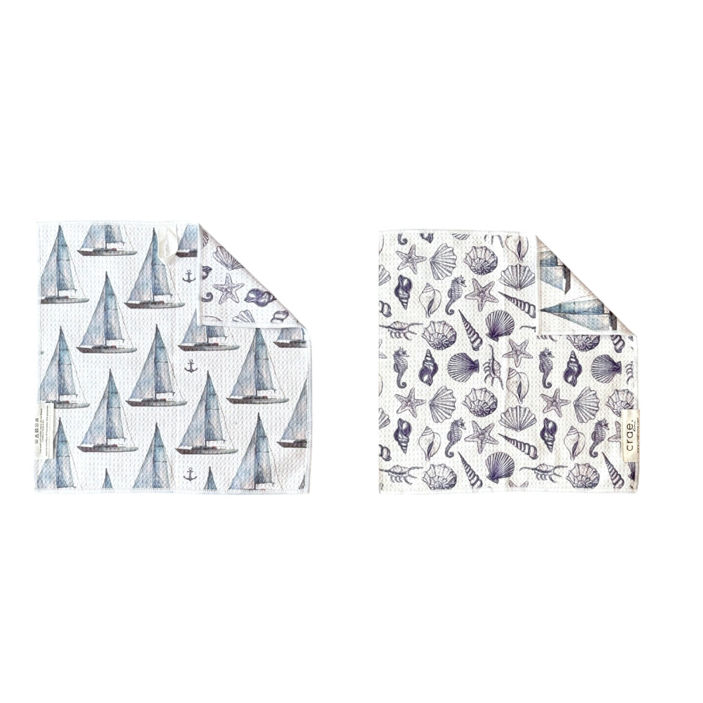 A double sided washcloth that  features a pattern of blue and gray sailboats, while the right napkin features a pattern of blue and gray seashells, starfish, and sea urchins.