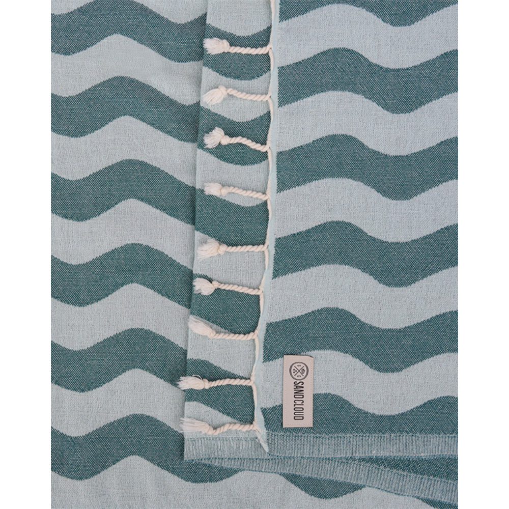 Colse up of a teal blue beach towel with blue wavy stripes and white tassel edge on a white background.