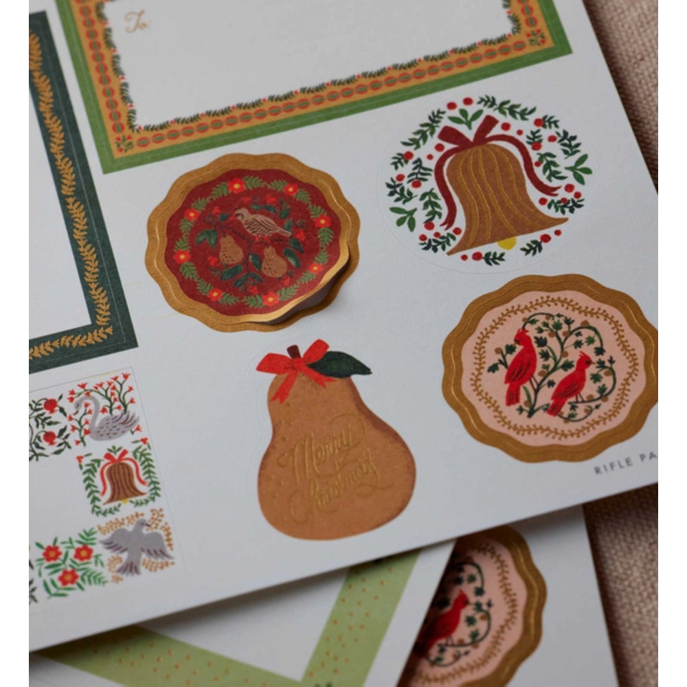 A close-up of a printed holiday card or stationery sheet featuring decorative Christmas illustrations, including a bell, pear, birds, and ornate borders in muted red, green, gold, and beige tones.