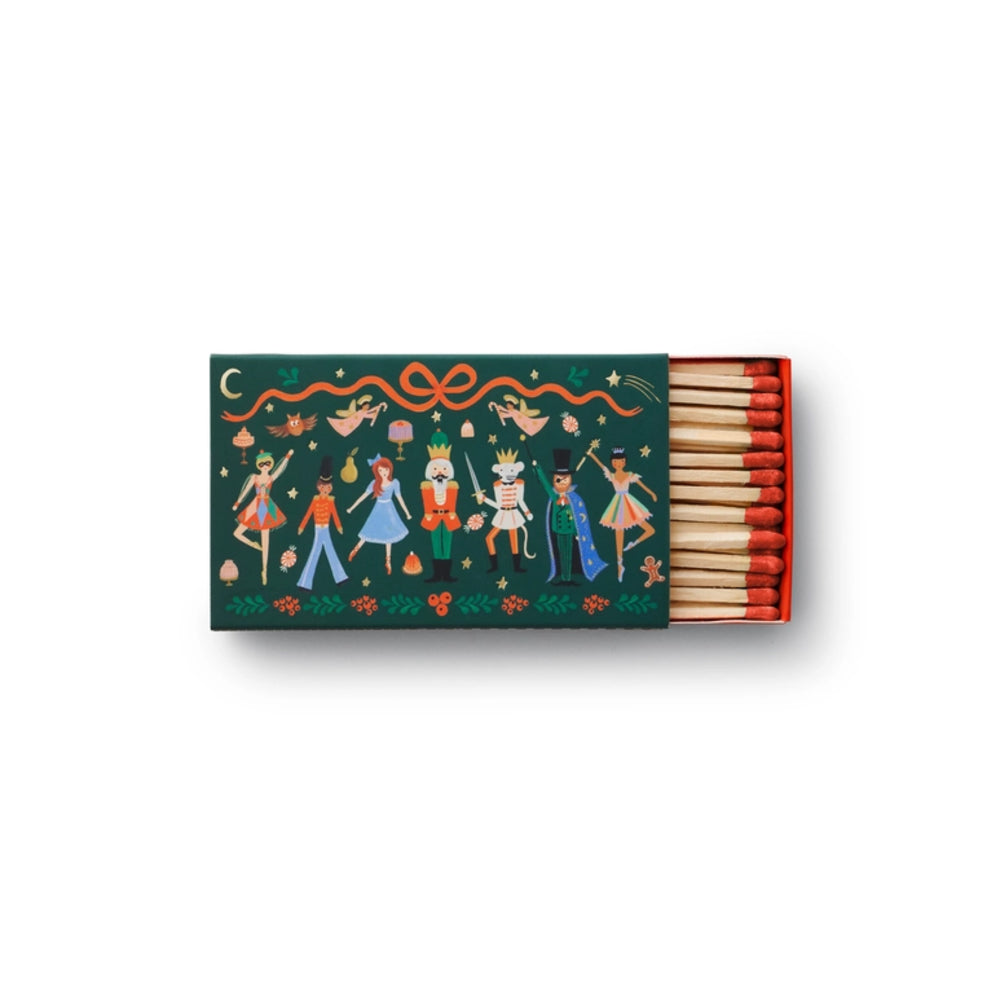 A decorative matchbox with a dark green cover featuring a colorful illustration of festive figures, a nutcracker, and holiday motifs, shown open with red-tipped matches on a white background.