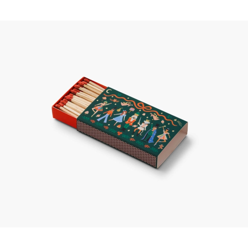 A decorative matchbox with a dark green cover featuring a colorful folk-art illustration of figures and animals, shown with wooden matches partially extended on a light gray background.
