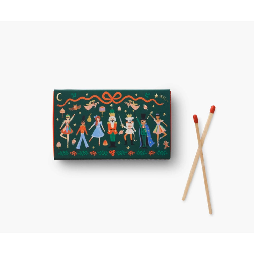 A dark green matchbox with a colorful folk-art illustration of figures, animals, and ornaments, shown next to two wooden matches with red tips on a clean white background.