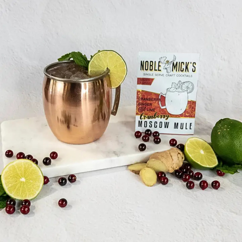 A copper Moscow mule mug with ice, mint, and lime garnish sits on a white marble board next to a box of Noble Mick's Single Serve Craft Cocktails Cranberry Ginger Moscow Mule mix, surrounded by cranberries, ginger, and lime slices on a white textured background.