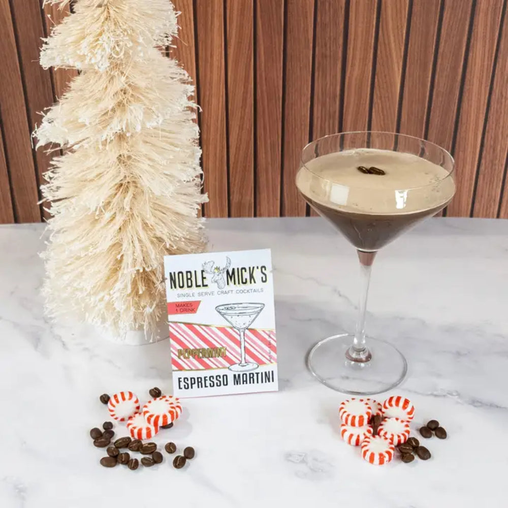 A styled product shot featuring a card that reads 'Noble & Mick's Espresso Martini' next to a prepared espresso martini in a coupe glass, with decorative peppermint candies, coffee beans, and a small beige tree against a wood-paneled background.