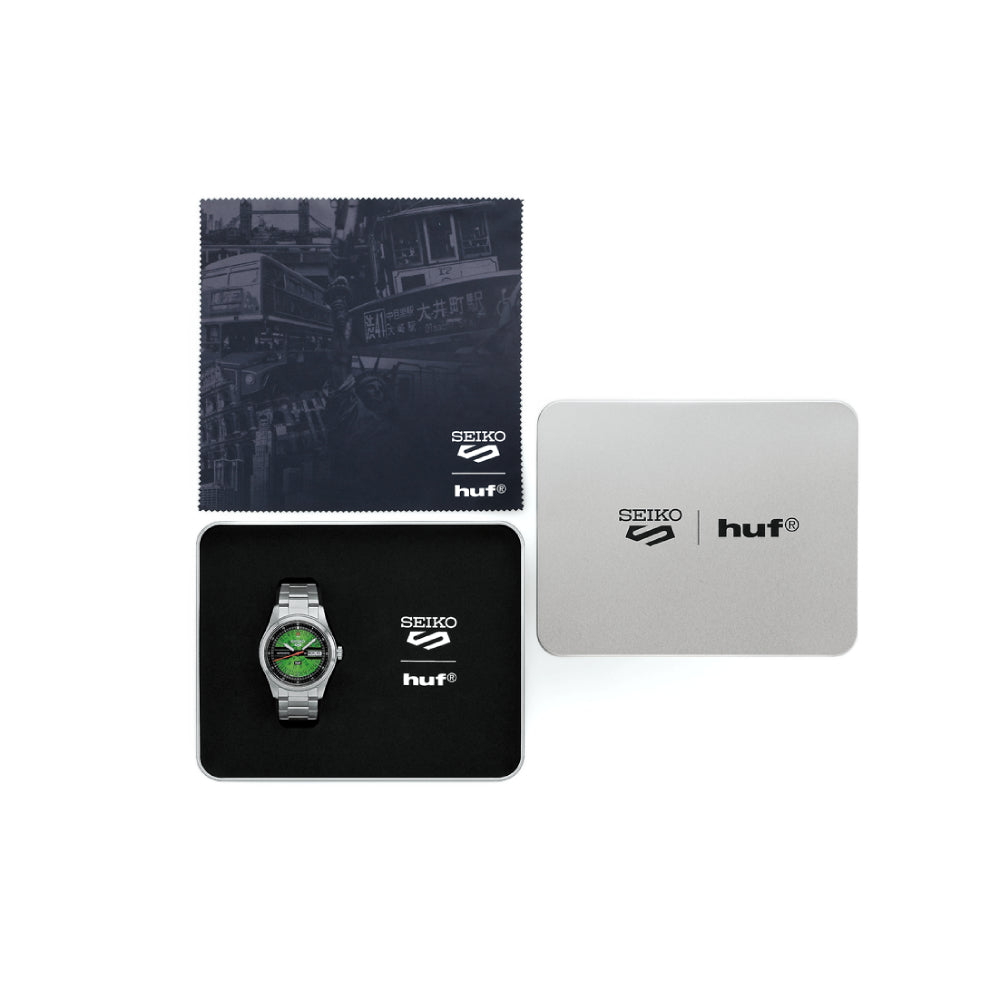 Steel Seiko watch with green "HUF" dial and included box and cleaning cloth on a white background