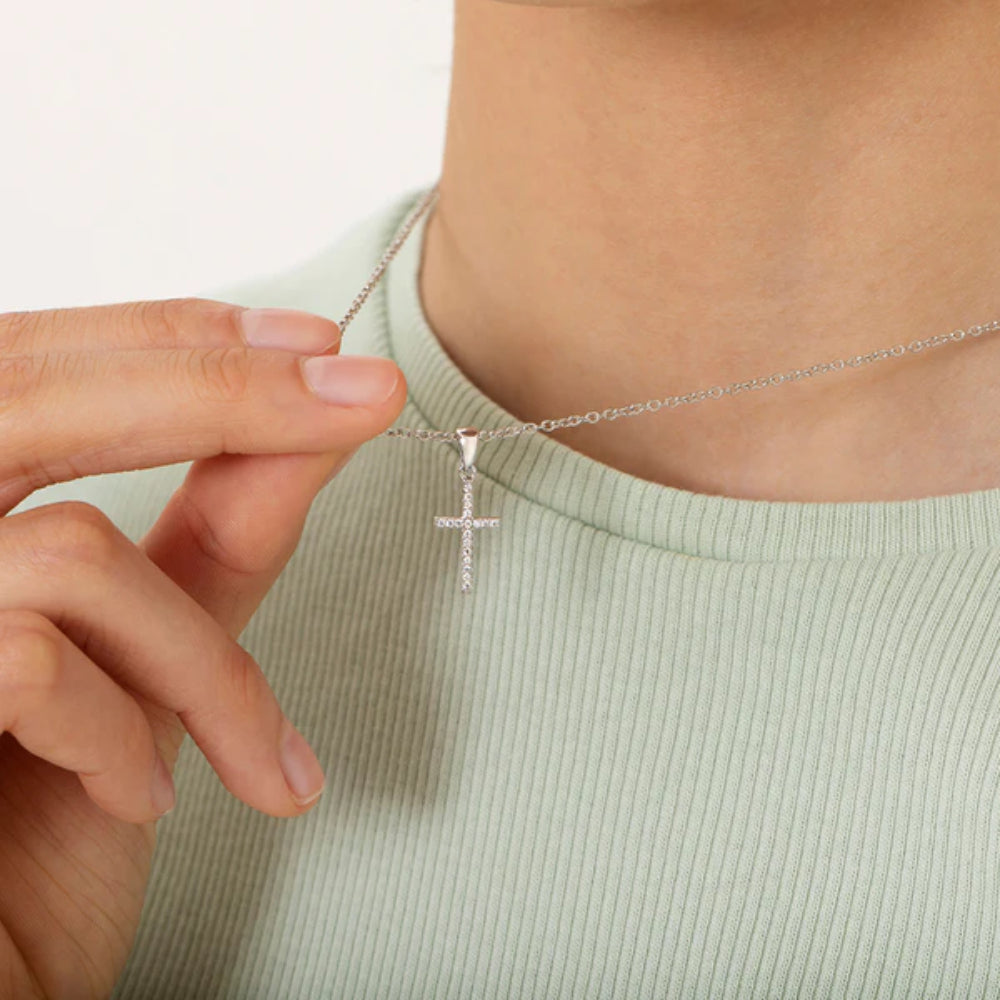 Person wearing a silver necklace with a cross pendant on a plain background