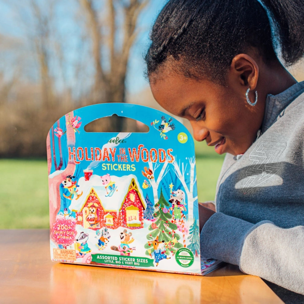 A child leans over a wooden table looking at a colorful 'Holiday in the Woods Stickers' package. The package features cartoon woodland characters and decorated houses on a bright blue background, set against a blurred outdoor park scene.