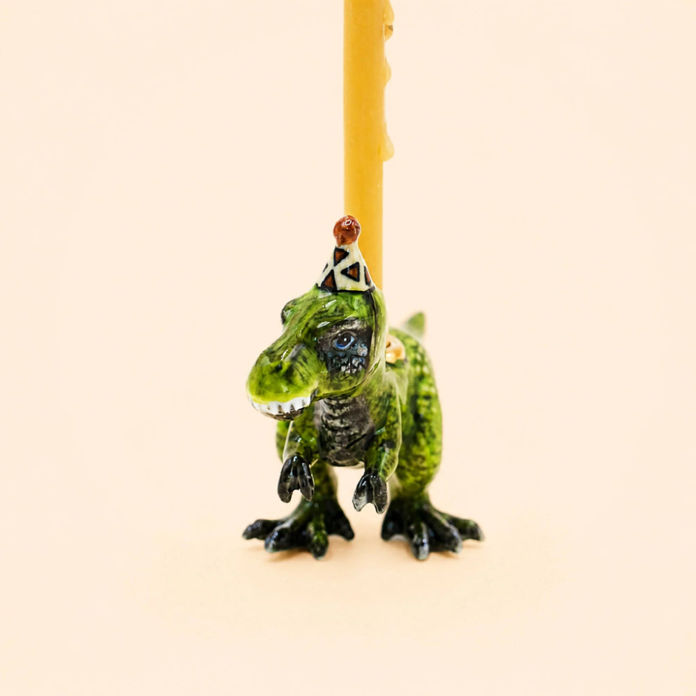 A small green dinosaur figurine wearing a party hat, positioned in front of a vertical yellow candle on a plain light beige background.