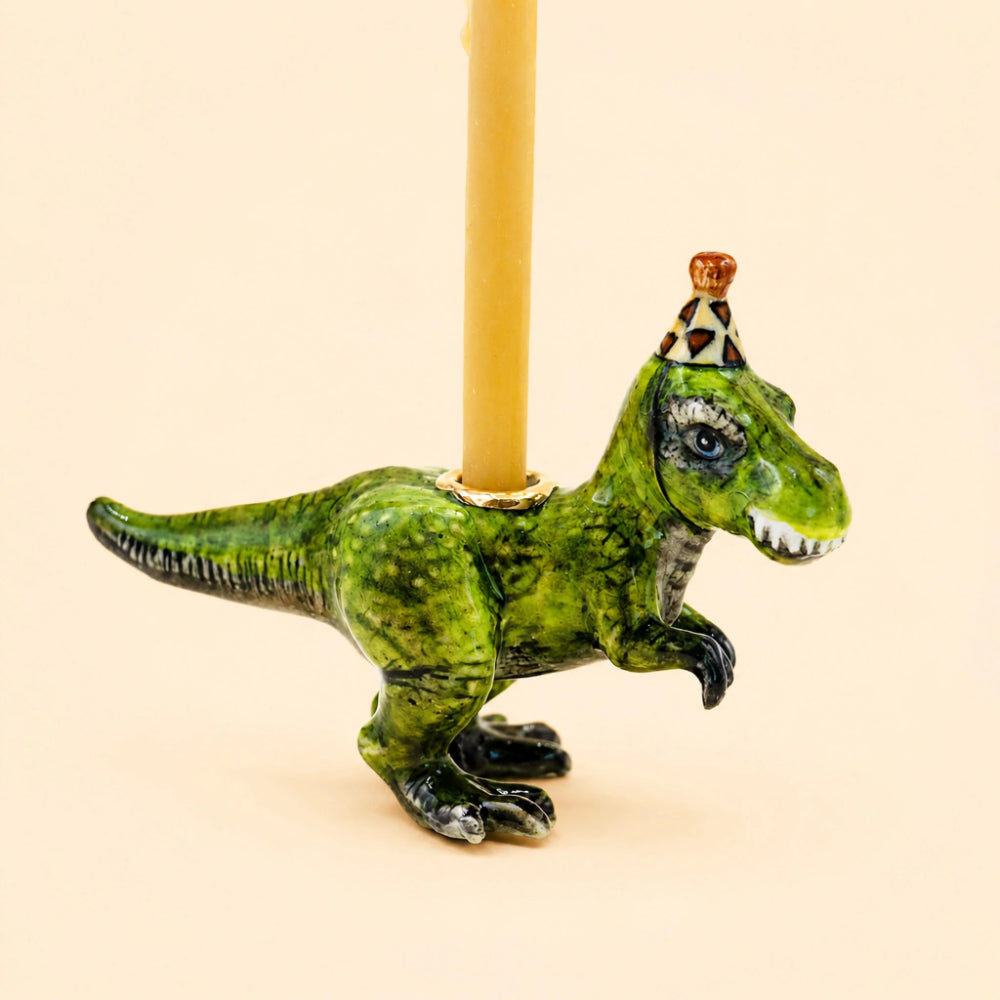 A green dinosaur figurine candle holder with a textured, mottled green and black body, wearing a small patterned party hat. A yellow candle is inserted into the top opening, set against a solid pale peach background.