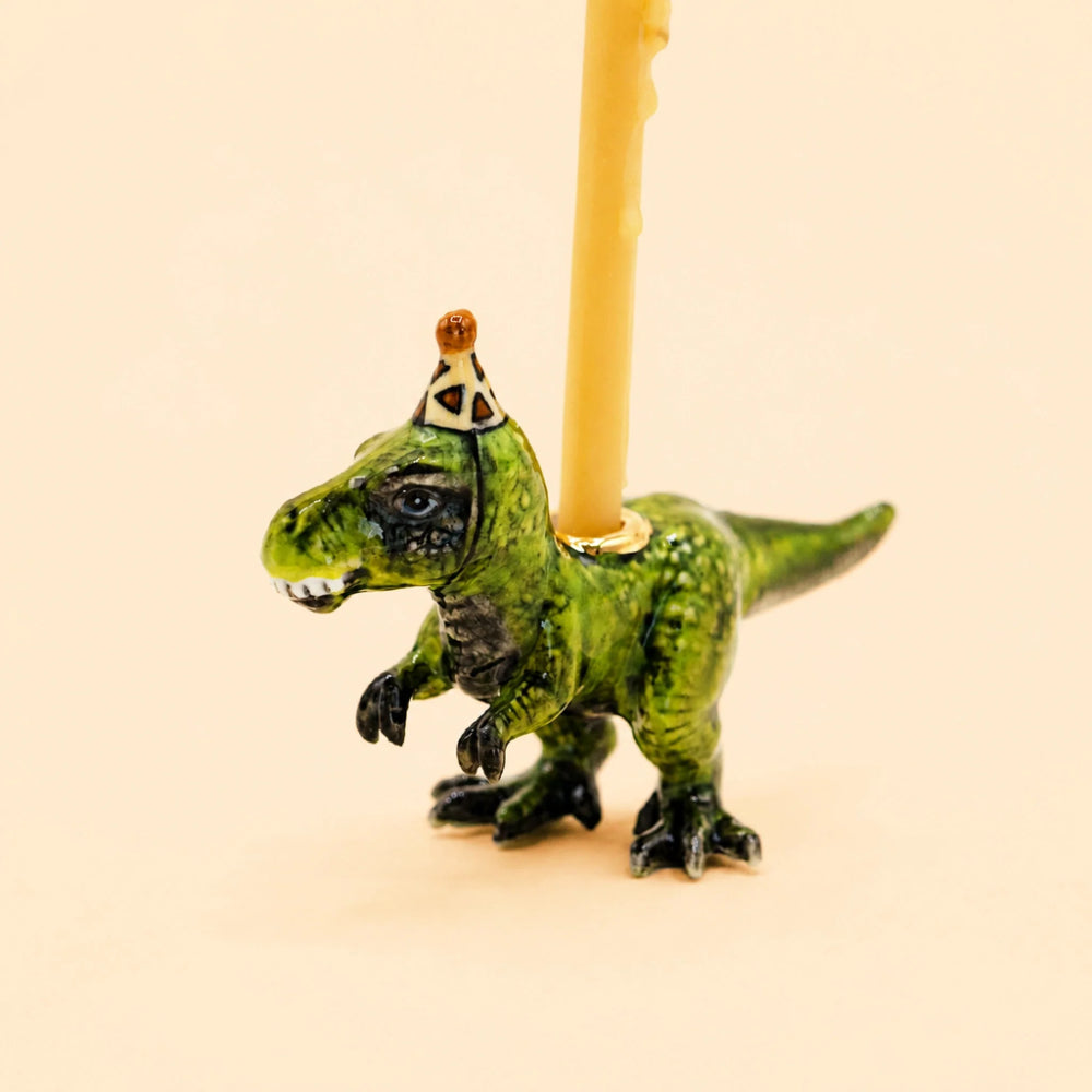 A small green dinosaur figurine with a gold crown, holding a yellow candle inserted into its back, set against a solid pale peach background.
