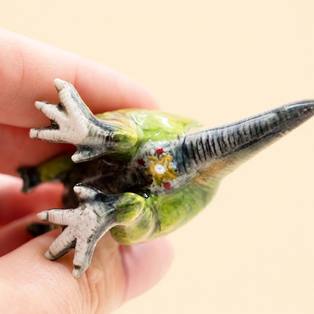 A small green and black dinosaur figurine with white claws, held in a person's hand against a plain beige background.