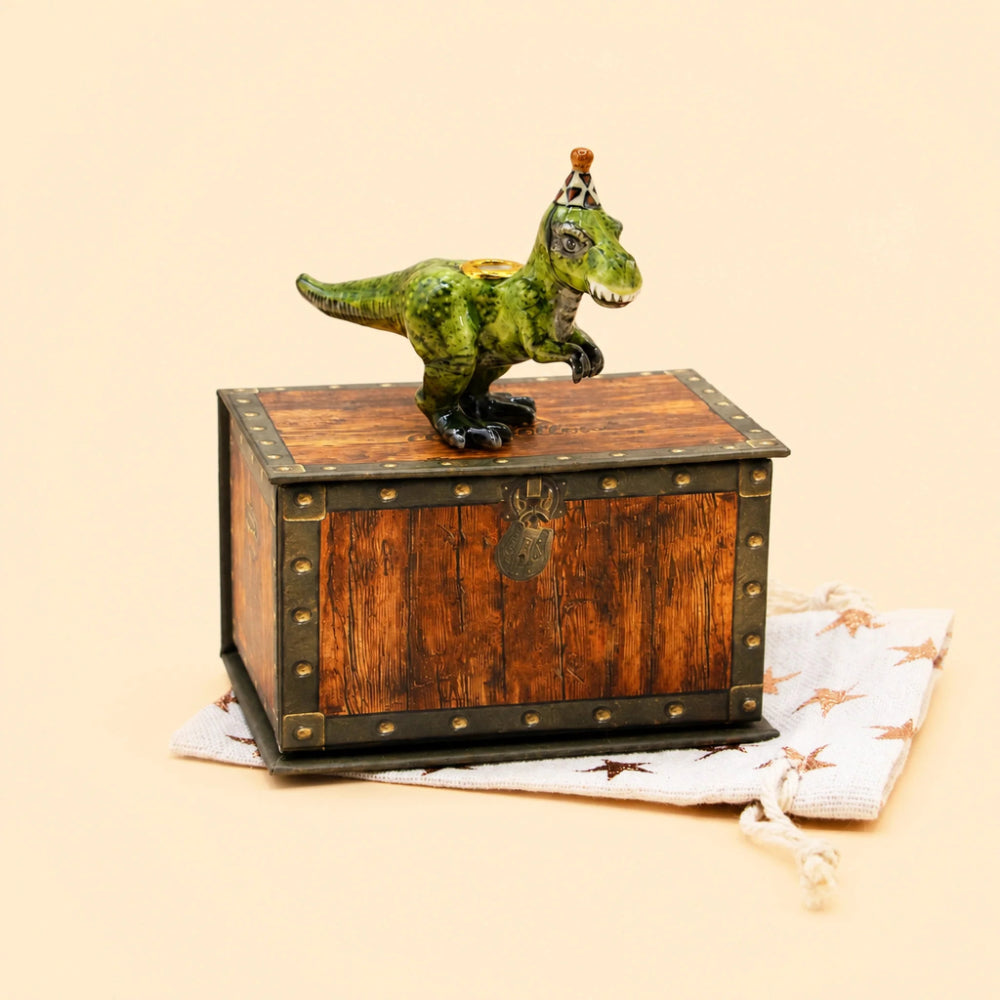 A small green dinosaur figurine with a miniature figure on its head stands atop a rustic brown wooden chest with metal corner studs. The chest rests on a white drawstring pouch printed with dinosaur silhouettes, against a plain warm beige background.
