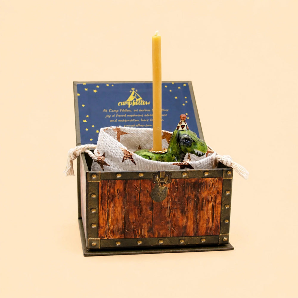 A miniature wooden treasure chest gift box with brass-style corner rivets, containing a small green dragon figurine wearing a crown, a white fabric lining, and a single yellow taper candle. The lid is open and displays a blue card with gold text and stars.