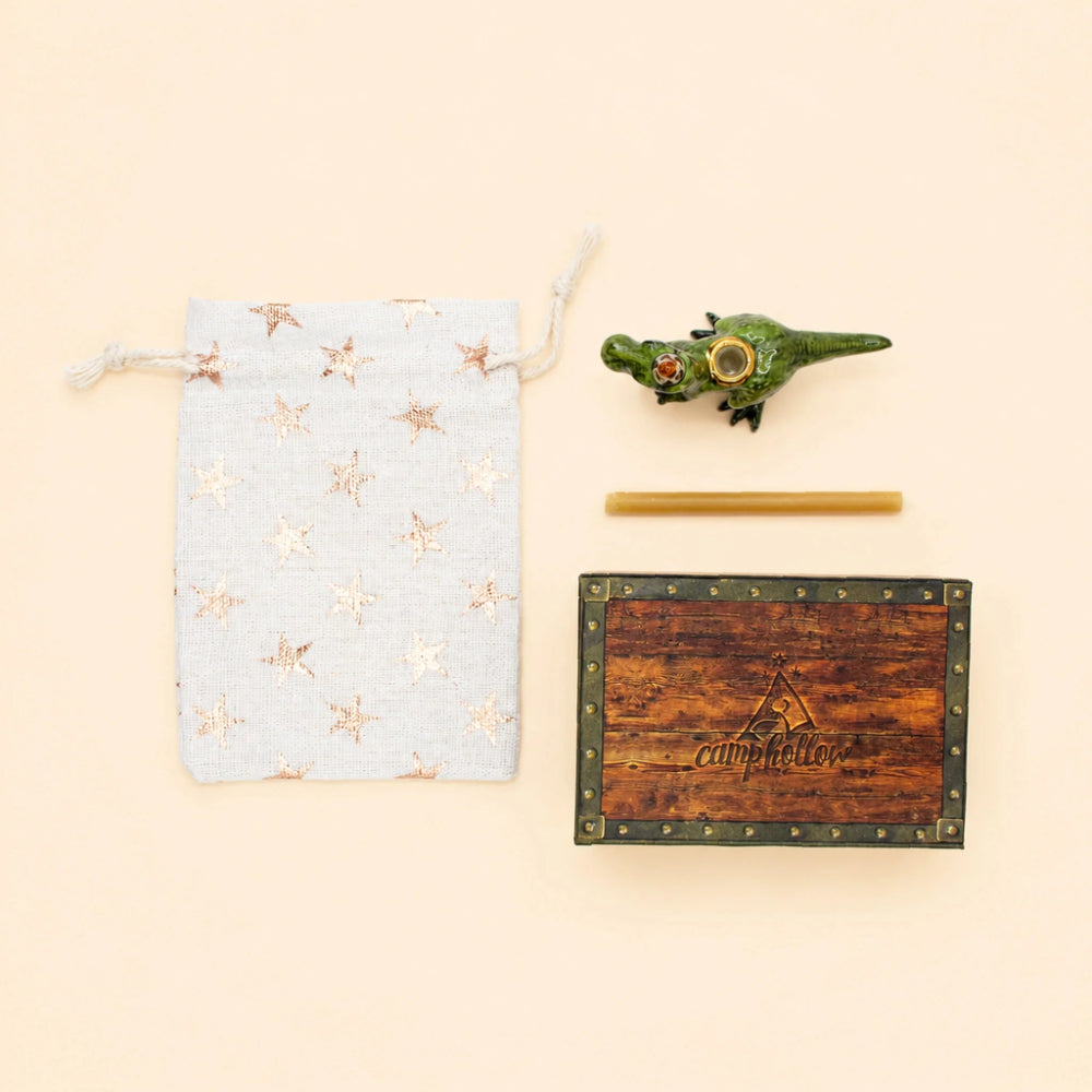 A flat-lay product shot featuring a light blue drawstring pouch with metallic gold star patterns, a small green crocodile-shaped candle holder, a wooden stick, and a wooden box with metal trim and the brand name 'Empshelter' embossed on the front, all arranged on a solid pale beige background.