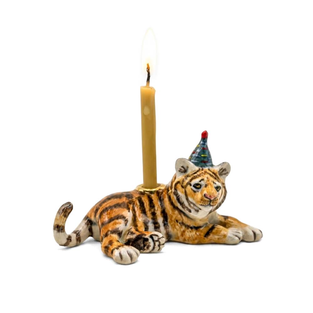 A ceramic tiger figurine candle holder on a white background, featuring a lit candle inserted into its back and a small party hat on its head.